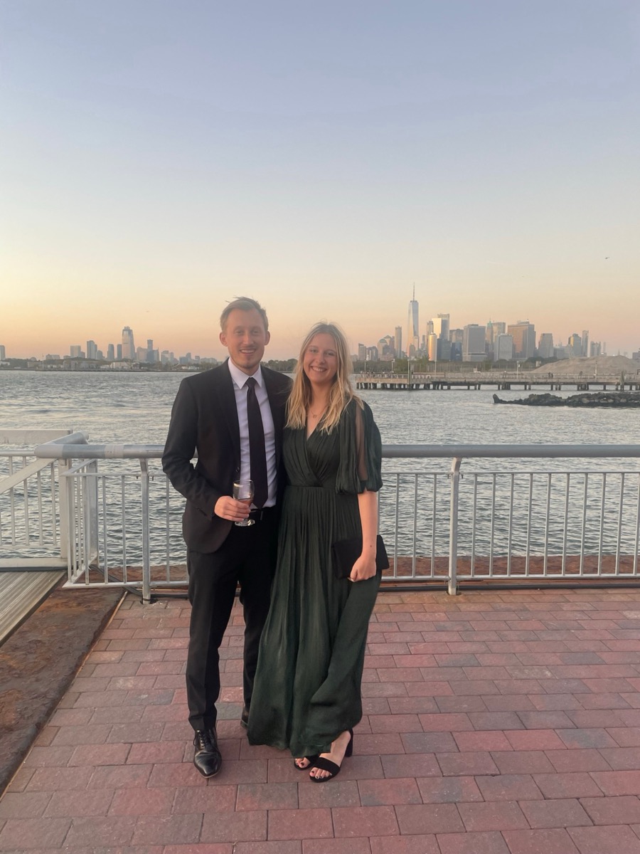 Patricia and Nikolas dressed up on a waterfront terrace with the Manhattan skyline at sunset